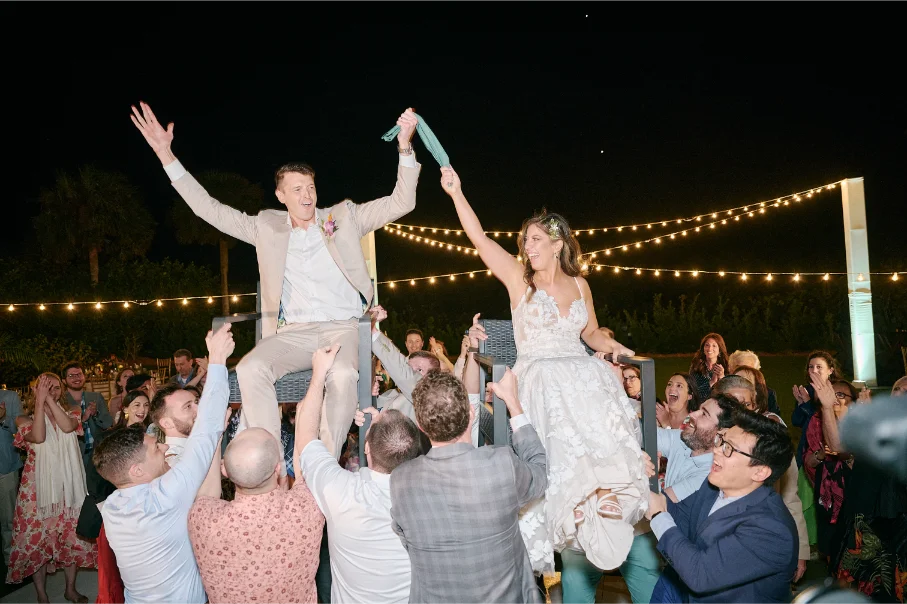 Bride and groom are crowd surfing on chairs, surrounded by lights and laughing people.